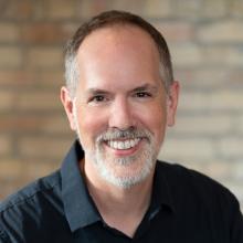 headshot of Dan Moriarty, wearing a dark blue shirt, in front of a brick wall