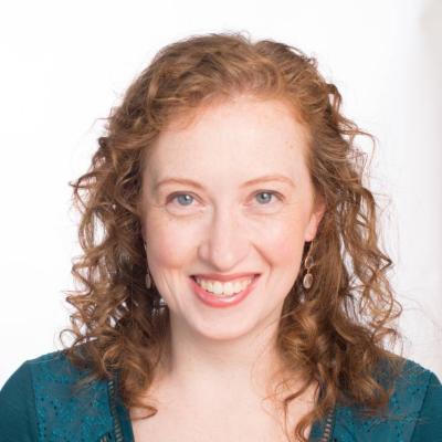 A woman with curly red hair wearing a deep green v-neck top smiling at the camera
